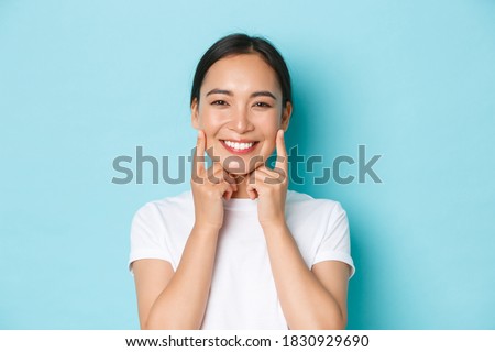 Close-up of beautiful asian girl looking pleased, smiling white teeth, touching cheeks, recommend skincare product, cosmetics for facial skin, standing light blue background