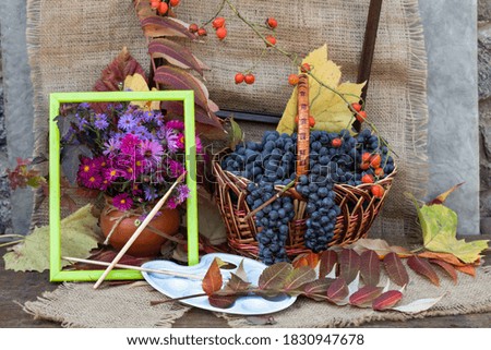 On a jute cloth, there is a basket of grapes, a clay pot with autumn flowers. Nearby are autumn leaves, an artist's palette, brushes and a picture frame.