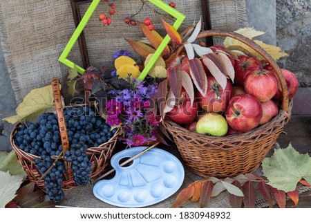 On a jute cloth, there is a basket of grapes and a basket of apples, a clay pot with autumn flowers. Nearby are autumn leaves, an artist's palette, brushes and picture frames.
