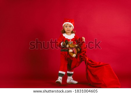 Cute little girl with her reindeer plush toy and Santa Claus sack against red background. Merry Cristmas and Happy New Year. Holiday and beauty concept
