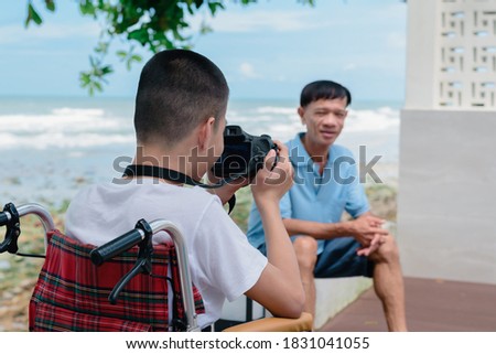 Asian special child on wheelchair is holding camera take a photo father at the sea beach, Boy happiness in holidays with parent in family time on the travel, Lifestyle of happy disabled kid concept.