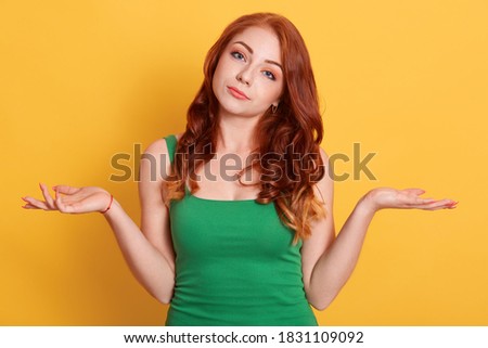 Photo of doubtful European woman faces dilemma, spreads hands sideways, looks at camera, has difficult decision, dressed casually stands over yellow wall.
