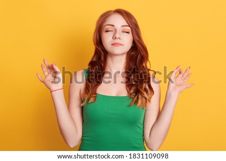 Red haired peaceful Caucasian woman in green t shirt meditates indoor against yellow wall, keeps hands in mudra gesture, has eyes closed, relaxes after long hours working, holds fingers in yoga sign
