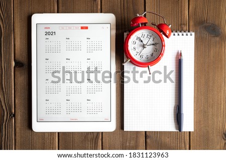 a tablet with an open calendar for 2021 year, a red alarm clock, and a spring notebook with a pen on a wooden boards table background Royalty-Free Stock Photo #1831123963