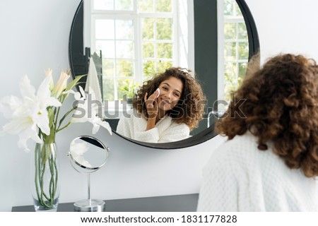 Natural beauty concept. Glad young afro american woman making morning routine, removing makeup, looking at mirror, holding cotton pad in hands near face and sitting in bathrobe at bathroom Royalty-Free Stock Photo #1831177828