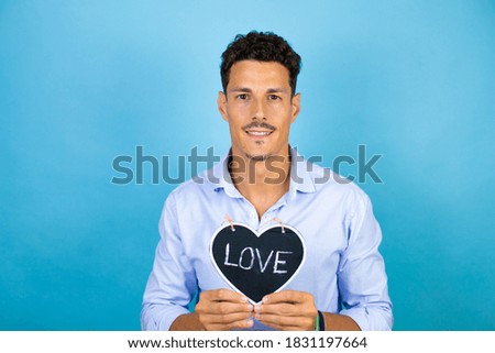 Young handsome man wearing blue shirt over isolated blue background smiling and holding heart blackboard with love word message