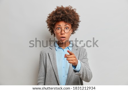 Shocked beautiful curly haired successful female entrepreneur points at you with stunned expression and presents something to colleagues comes on formal meeting stands indoor against grey background