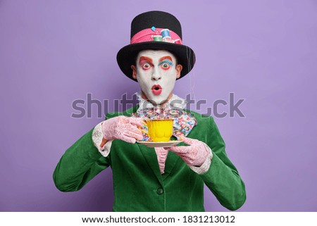 Mad tea party. Crazy shocked man hatter wears large top hat oversized bowtie lace gloves and green jacket drinks hot beverage has image of fictional character celebrates halloween stands indoor Royalty-Free Stock Photo #1831213012