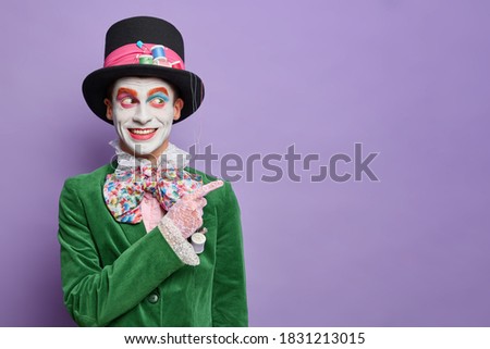 Smiling man dresses for carnival party has image of hatter from wonderland indicates away on blank space wears halloween costume and bright makeup isolated on purple background. Street performer