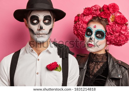 Close up shot of loving couple with spooky makeup pose during day of dead enjoy halloween party stand against rosy background wear mexican attire. Dead woman and man celebrate holiday in Mexico