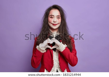 Beautiful smiling woman with fearful makeup pale ghost face and bloody scars makes heart gesture and expresses love being on halloween party isolated over purple background. Mystical character