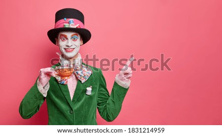 Positive man has image of hatter enjoys tea ceremony poses with cup in carnival costume and points away on blank space for your promotion prepares for halloween. Holiday masqeurade performer