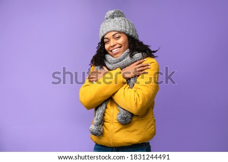 Winter Clothing. Happy African American Woman Wearing Warm Jacket, Cap And Scarf Standing Posing In Studio Over Purple Background, Looking At Camera. Cold Weather Clothes Fashion Concept