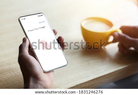 Biometric Authentication Concept. Over The Shoulder View Of Person Holding Mobile Phone In Hand Showing Application For Fingerprint Scanning With Thumbprint Icon On Device Screen, Drinking Coffee