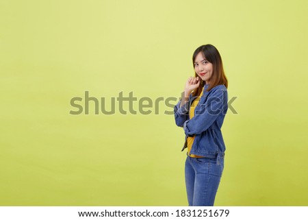 A portrait of a young, cute and athletic Thai Asian girl in a studio against a plain yellow background. Woman with crossed arms wearing yellow t-shirt jeans jacket and simple jeans