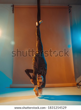 young woman in black costume is making aerial trick upside down on the beige background in photo studio in warm and cold lights, sport concept, free space