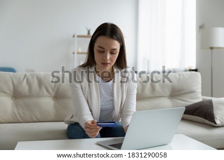 Young Caucasian woman sit on sofa in living room shopping online on laptop with credit card. Millennial female make internet payment on computer, enter bank details, use secure web service system.