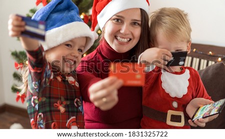 Xmas, winter, new year, Celebration, family concept - joyful mom doing shopping online with two small funny kids sit near Christmas tree hold discount cards and phone in hands, gifts for relatives