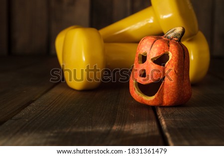 Small ceramic Halloween pumpkin with two yellow dumbbells. Healthy fitness lifestyle autumn composition on wooden background.