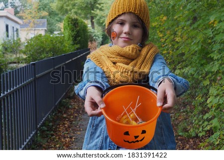 A boy in an orange  knitted hat, scarf, with a pumpkin make-up on his cheek holds a jack-o-lantern bucket with candies in it. Halloween theme