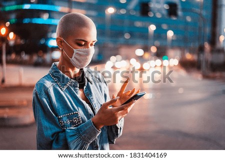 Pretty girl with stylish clothes holding smartphone outdoors in the evening, illuminated city on background - Concepts about technology, communication and lifestyle during covid-19 pandemic