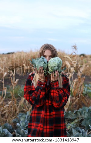 A blonde girl in a red plaid shirt, holds two cabbages in her hands against her head, and hides a little.