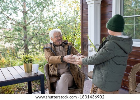 Joyful grandfather in warm casualwear sitting in armchair by small table and passing birthday surprise in giftbox to his grandson by house
