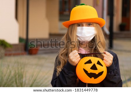 Little girl in Halloween costume with pumpkin bucket near the house. Candy or life, child wearing white protective face mask outdoors. Celebrating Halloween during COVID-19.