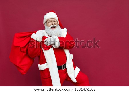 Happy old funny Santa Claus, Saint Nicholas wearing costume holding sack bag with Merry Christmas presents walking delivering xmas gifts standing isolated on red background. Xmas delivery concept. Royalty-Free Stock Photo #1831476502