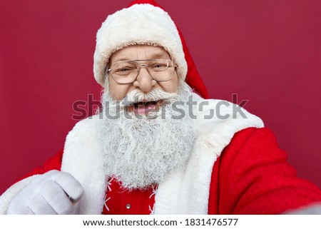 Happy old bearded Santa Claus wearing costume holding phone in hand taking selfie, video calling, recording video Merry Christmas greeting or shooting vlog isolated on red background, camera view.