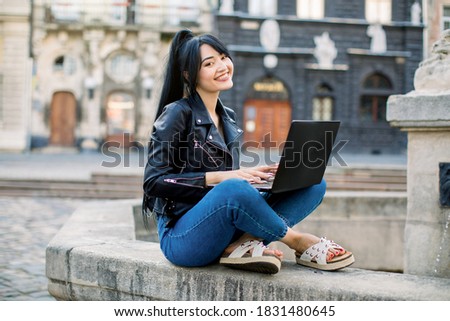 Free Wi-Fi in the city, freelance concept. Smiling young mixed raced Asian woman with black ponytail hair sitting on fountain and texting message on laptop, checking news in social networks.