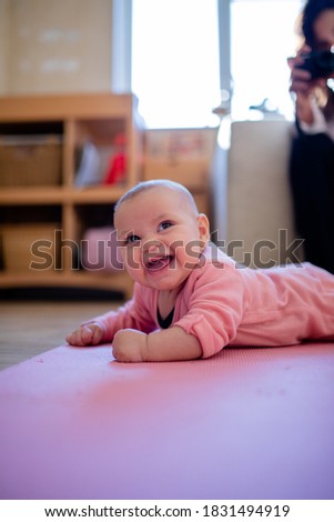 Portrait style picture of a smiling baby in a pink sleepsuit lying face down on a pink rug with a blurry person photographing her in the background