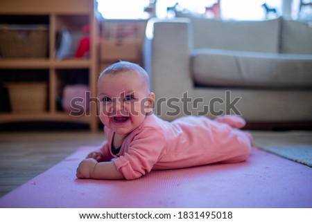 Picture of a smiling baby in a pink sleepsuit lying face down on a pink rug with a couch and a window as background
