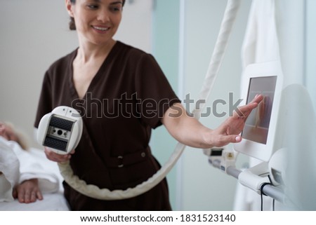 Cropped photo of smiling cosmetologist pressing the screen of special modern equipment in beauty clinic