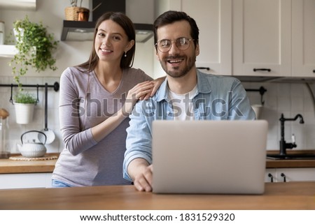 Portrait of happy millennial married family couple sitting at kitchen table with computer, enjoying relaxed weekend morning time web surfing information, young people and modern technology concept.