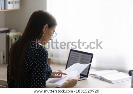 Back rear view focused young woman in eyeglasses reading paper document, checking electronic report on computer. Concentrated smart ambitious businesswoman preparing research, freelance work. Royalty-Free Stock Photo #1831529920