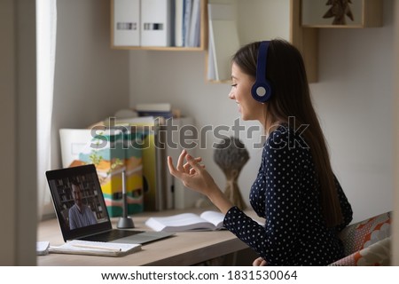 Side view happy young caucasian female student in earphones involved in interesting online discussion with teacher, holding computer video call from home, e-learning distant education concept. Royalty-Free Stock Photo #1831530064