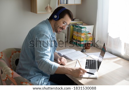 Happy young smart guy in glasses and wireless earphones looking at copybook notes, listening to educational lecture, enjoying study on online courses remotely at home office, e-learning concept. Royalty-Free Stock Photo #1831530088