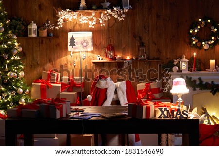 Santa Claus costume and hat hanging on chair at table with Merry Christmas decor gifts presents on holiday eve in cozy Santa home workshop interior late in night with light on xmas tree and fireplace. Royalty-Free Stock Photo #1831546690