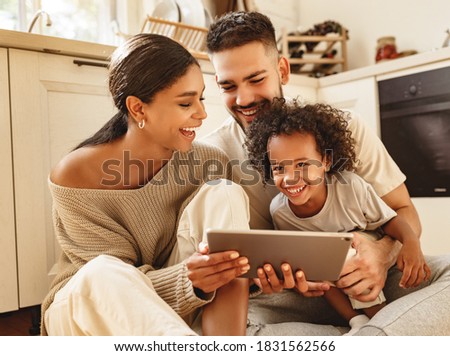 happy multi ethnic family: parents and little son laugh and watch funny video on a tablet while sitting on the kitchen floor at home
