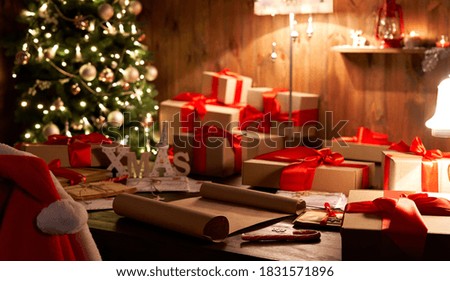 Santa Claus costume and hat hanging on chair at table with Merry Christmas wish list decor gifts presents on holiday eve in cozy Santa home workshop interior late in night with light on xmas tree. Royalty-Free Stock Photo #1831571896
