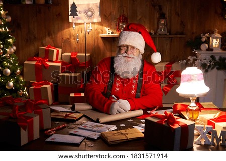 Portrait of happy old kind bearded Santa Claus wearing hat, glasses, looking at camera, working on Christmas eve sitting at cozy home table late with presents, tree and candles preparing for holidays. Royalty-Free Stock Photo #1831571914