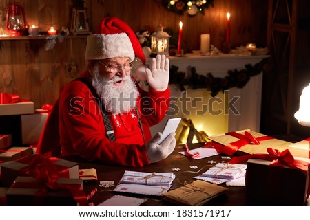 Happy Santa Claus holding smartphone video calling kid talking to child in virtual video online chat meeting sit at home table late with presents on xmas eve. Merry Christmas social distance concept. Royalty-Free Stock Photo #1831571917