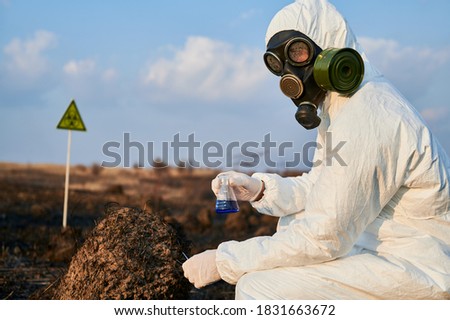 Scientist in protective suit, gas mask holding test tube with blue liquid while studying burnt grass and soil on scorched territory with biohazard sign. Concept of ecology, research and burned earth
