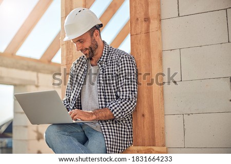 Handsome young man in a hard hat looking at laptop screen while sitting outside the construction site Royalty-Free Stock Photo #1831664233