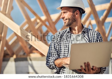 Waist-up photo of a brunette man in a hard hat holding a laptop and looking at the construction site