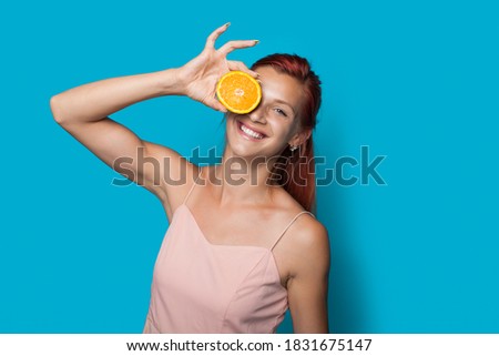 Red haired woman is covering her eye with a sliced lemon posing on a blue studio wall