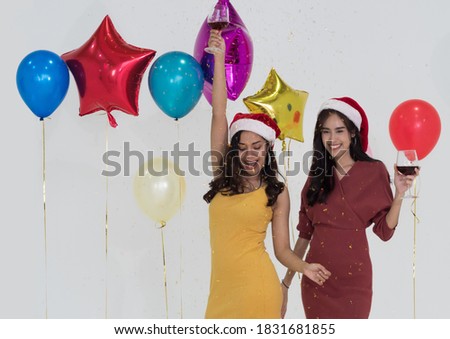 Two happy Asian women wearing Christmas party hat holding red wine glasses dancing with colorful balloons as background.