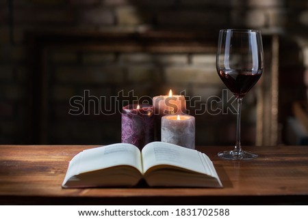Glass of red wine, open book with candles in the background on wooden table. Warm, dark colors.