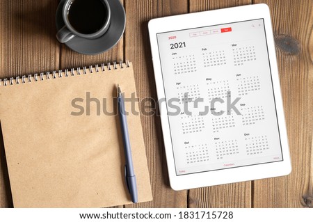 a tablet with an open calendar for 2021 year, a cup of coffee and a craft paper notebook on a wooden boards table background Royalty-Free Stock Photo #1831715728